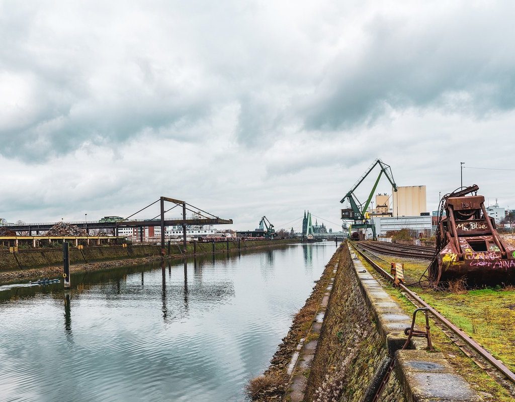 Blick über den Deutzer Hafen in Köln an einem bewölkten Tag: Ein ruhiger Hafenkanal verläuft mittig durchs Bild, links industrielle Anlagen und Schrottlager, rechts alte Gleise mit einem rostigen Greifbagger. Im Hintergrund sind Kräne, Industriegebäude und die Kölner Skyline mit Kirchtürmen zu sehen.