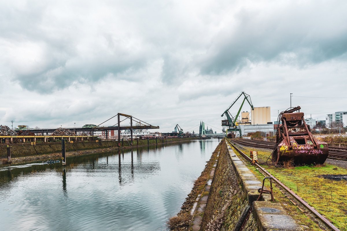 Blick über den Deutzer Hafen in Köln an einem bewölkten Tag: Ein ruhiger Hafenkanal verläuft mittig durchs Bild, links industrielle Anlagen und Schrottlager, rechts alte Gleise mit einem rostigen Greifbagger. Im Hintergrund sind Kräne, Industriegebäude und die Kölner Skyline mit Kirchtürmen zu sehen.