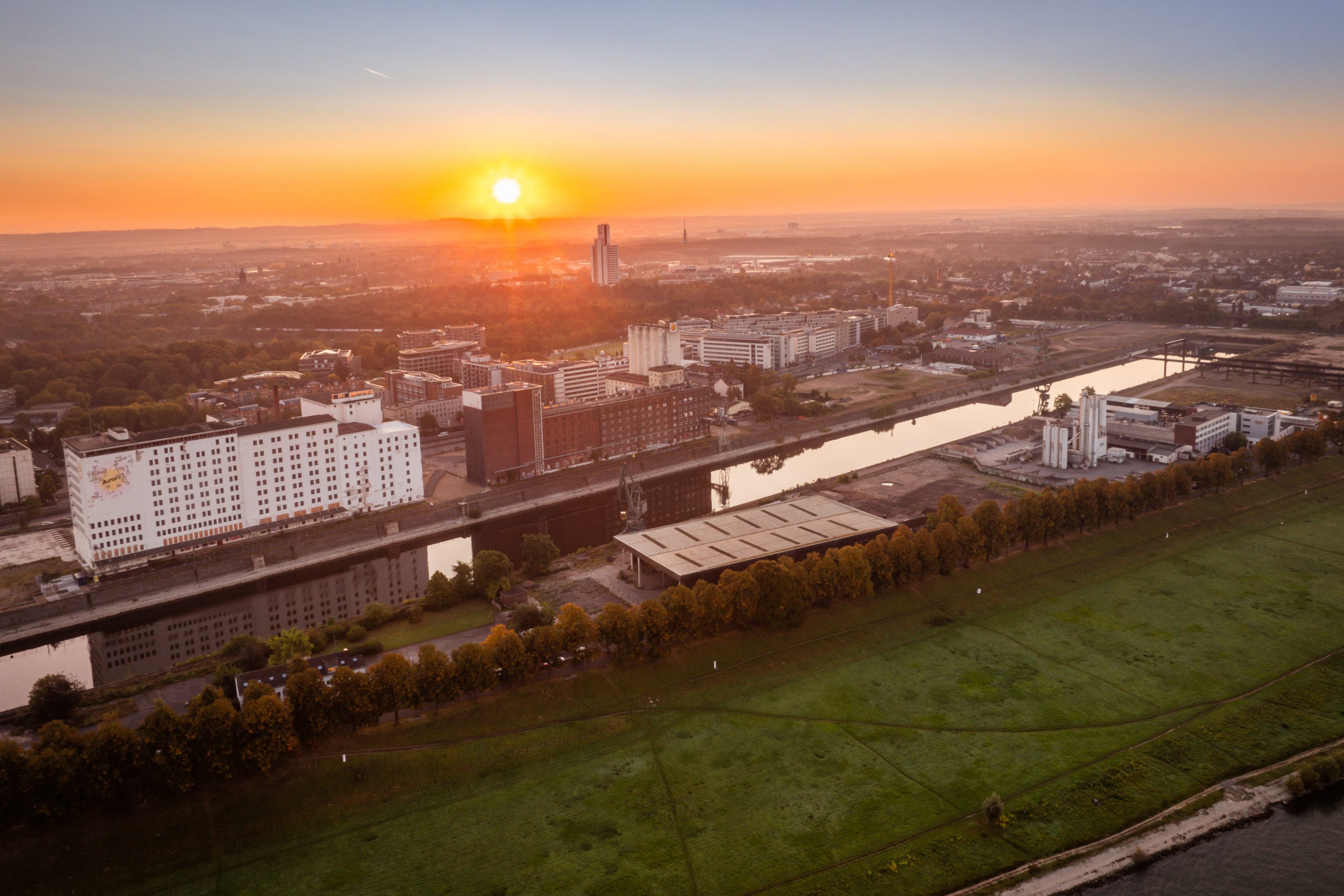 Luftaufnahme des Deutzer Hafens in Köln bei Sonnenaufgang: Der Hafenkanal verläuft durch das ehemalige Industriegebiet mit Mühlengebäuden, Silos und Gewerbebauten. Im Vordergrund liegt die Rheinwiese, im Hintergrund erstreckt sich die Stadtlandschaft im warmen Morgenlicht.