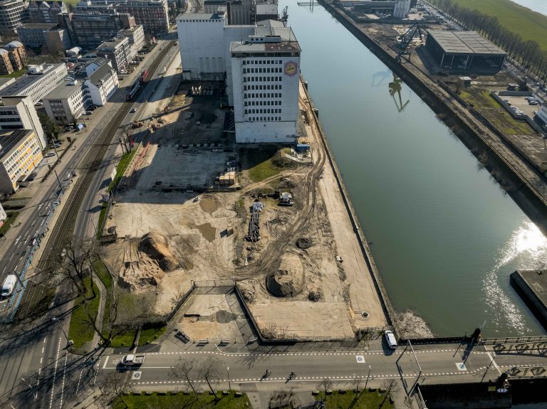 Luftaufnahme des Baufelds am westlichen Ufer des Hafenbeckens. Die weiße Ellmühle steht direkt am Wasser, davor ein geräumtes Baufeld mit Erdaufschüttungen und Baumaterial. Links die Straßenbahntrasse, rechts das spiegelglatte Hafenbecken mit der Poller Wiese. Im Vordergrund die Drehbrücke.