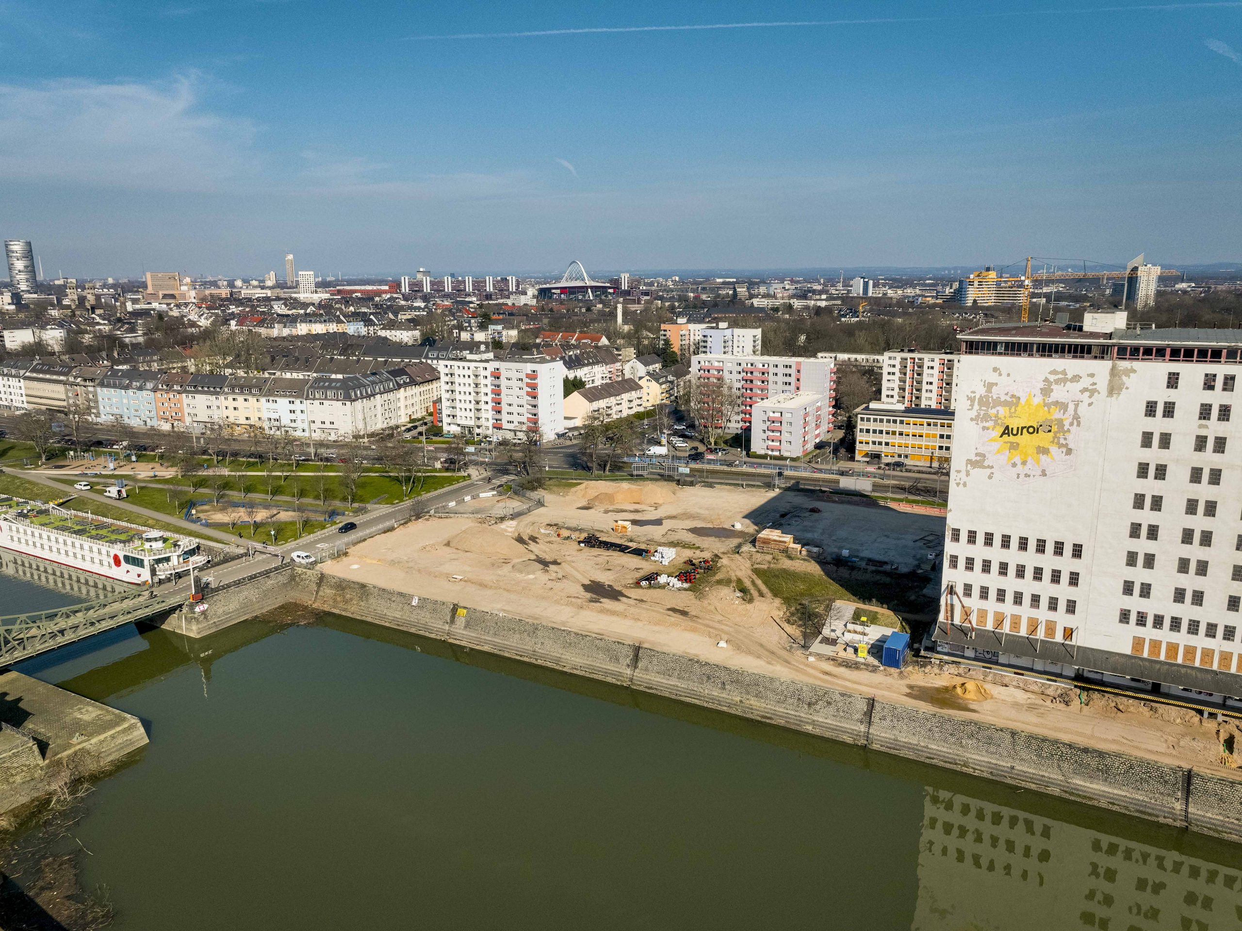 Luftaufnahme des nördlichen Baufelds am Deutzer Hafen bei blauem Himmel. Rechts die weiße Fassade der Ellmühle mit dem gut lesbaren Schriftzug „Aurora". Das geräumte Baufeld mit Baumaterial, einem blauen Baucontainer und Kran nimmt die Bildmitte ein. Links liegt ein Flusskreuzfahrtschiff an der Drehbrücke. Im Hintergrund die Kölner Skyline mit Dom und Colonius.