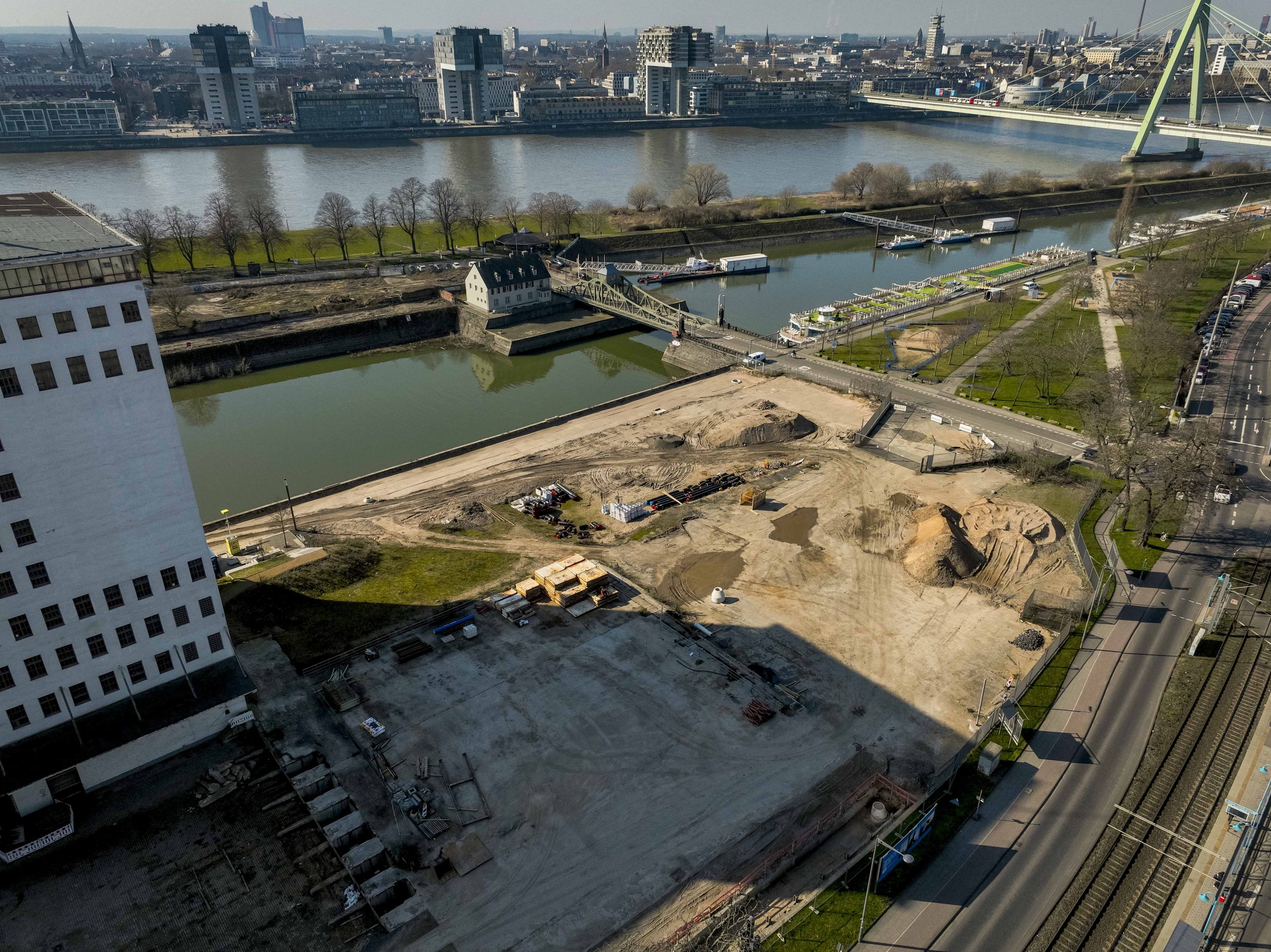 Drohnenaufnahme mit Blick vom Baufeld am Deutzer Hafen Richtung Kölner Innenstadt. Im Vordergrund das geräumte Baugelände mit Erdaufschüttungen. In der Bildmitte das Brückenwärterhäuschen der Drehbrücke und die Hafeneinfahrt. Dahinter Flusskreuzfahrtschiffe am Rheinufer. Im Hintergrund der Rhein mit der Severinsbrücke und die Kölner Stadtsilhouette.