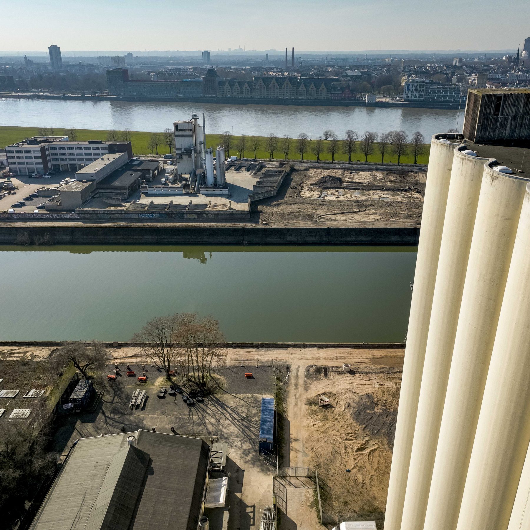 Drohnenaufnahme vom Dach der Ellmühlen-Silos am Deutzer Hafen. Im rechten Vordergrund die wuchtigen weißen Siloröhren der Ellmühle. Links daneben das Baufeld mit Baumaterial und einer alten Lagerhalle. Das Hafenbecken trennt das Bild von der Hafenzunge auf der anderen Seite. Im Hintergrund der Rhein mit dem Kölner Rheinpanorama und dem Rheinauhafen.