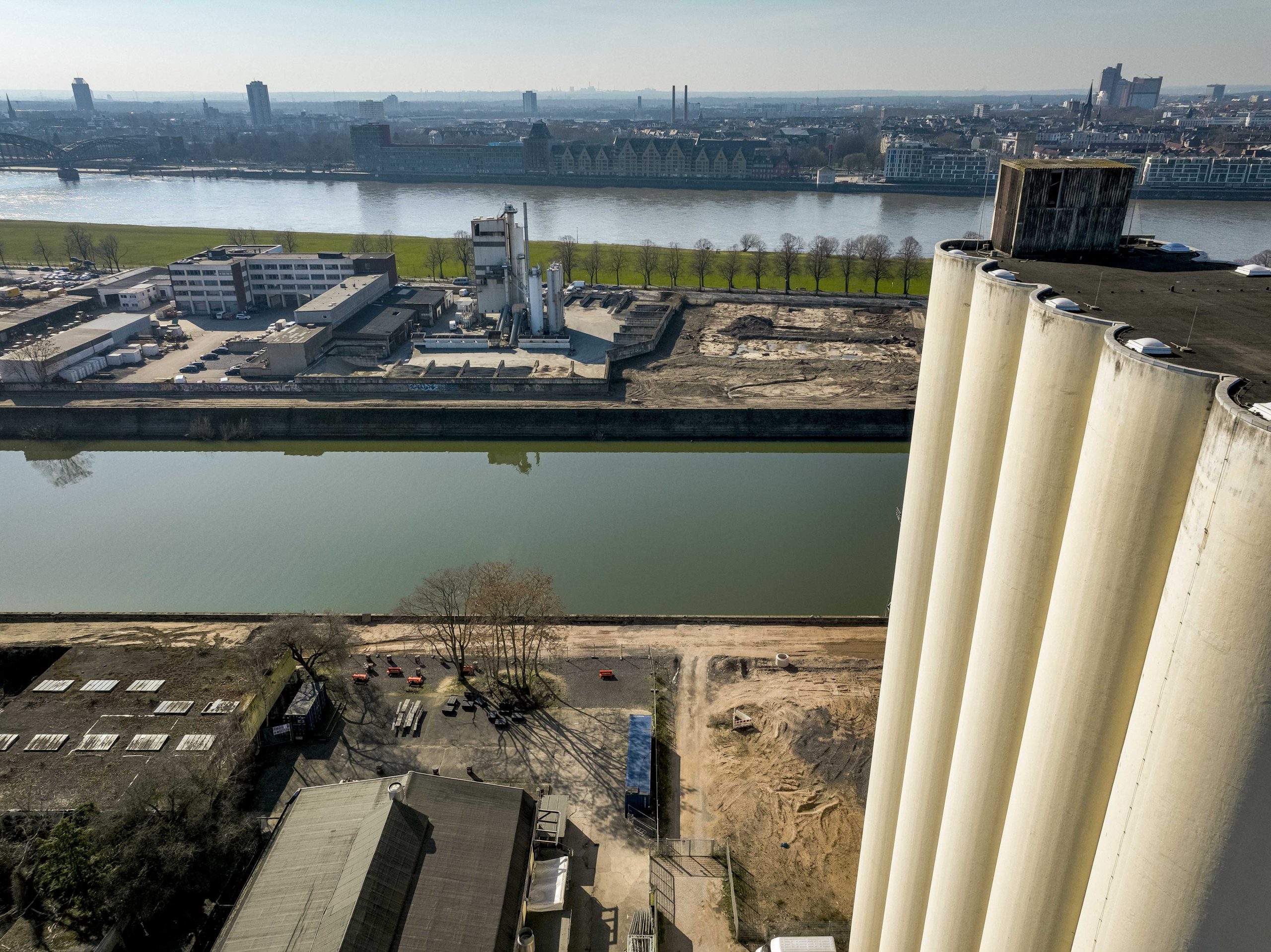 Drohnenaufnahme vom Dach der Ellmühlen-Silos am Deutzer Hafen. Im rechten Vordergrund die wuchtigen weißen Siloröhren der Ellmühle. Links daneben das Baufeld mit Baumaterial und einer alten Lagerhalle. Das Hafenbecken trennt das Bild von der Hafenzunge auf der anderen Seite. Im Hintergrund der Rhein mit dem Kölner Rheinpanorama und dem Rheinauhafen.