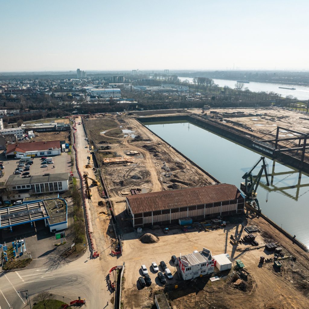 Drohnenaufnahme des südlichen Baufelds am Deutzer Hafen mit Blick auf das Hafenbecken. Im Vordergrund eine Lagerhalle und Baucontainer sowie ein grüner Hafenkran. Links eine Tankstelle und Gewerbegebäude an der Straße. Im Hintergrund das Hafenbecken mit dem Kunstwerk „See Things From The Other Side" sowie dem Portalkran. Rechts die Rodenkirchener Brücke.