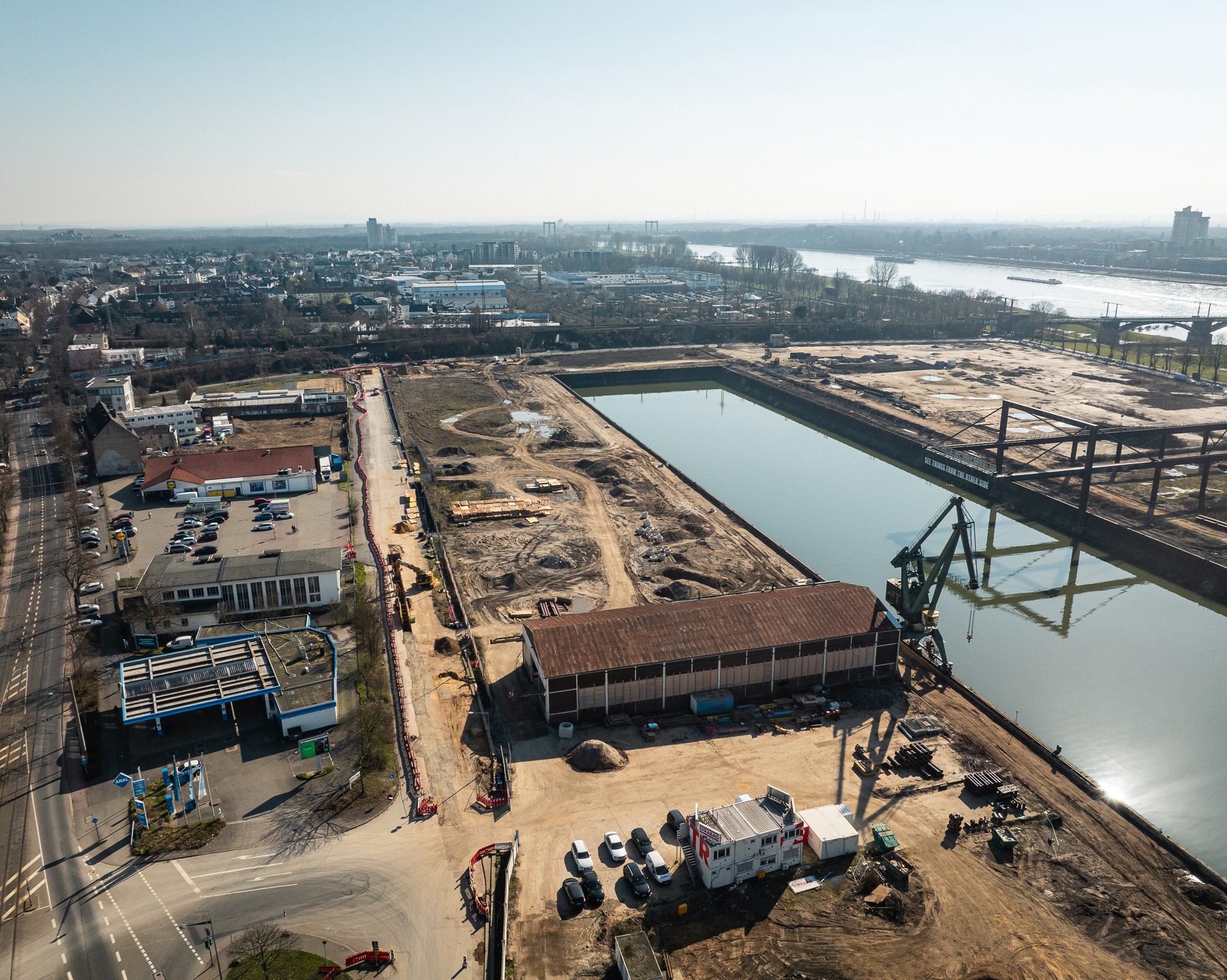 Drohnenaufnahme des südlichen Baufelds am Deutzer Hafen mit Blick auf das Hafenbecken. Im Vordergrund eine Lagerhalle und Baucontainer sowie ein grüner Hafenkran. Links eine Tankstelle und Gewerbegebäude an der Straße. Im Hintergrund das Hafenbecken mit dem Kunstwerk „See Things From The Other Side" sowie dem Portalkran. Rechts die Rodenkirchener Brücke.