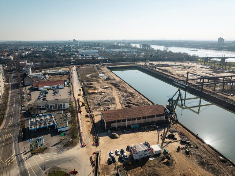 Drohnenaufnahme des südlichen Baufelds am Deutzer Hafen mit Blick auf das Hafenbecken. Im Vordergrund eine Lagerhalle und Baucontainer sowie ein grüner Hafenkran. Links eine Tankstelle und Gewerbegebäude an der Straße. Im Hintergrund das Hafenbecken mit dem Kunstwerk „See Things From The Other Side" sowie dem Portalkran. Rechts die Rodenkirchener Brücke.