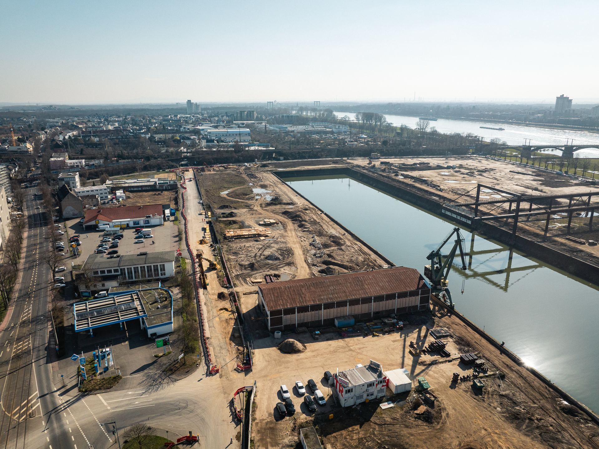 Drohnenaufnahme des südlichen Baufelds am Deutzer Hafen mit Blick auf das Hafenbecken. Im Vordergrund eine Lagerhalle und Baucontainer sowie ein grüner Hafenkran. Links eine Tankstelle und Gewerbegebäude an der Straße. Im Hintergrund das Hafenbecken mit dem Kunstwerk „See Things From The Other Side" sowie dem Portalkran. Rechts die Rodenkirchener Brücke.