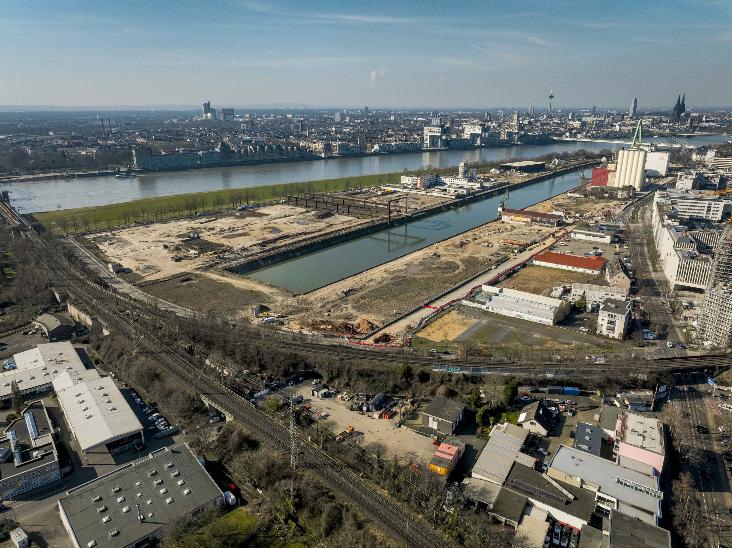 Weiträumige Luftaufnahme des gesamten Deutzer Hafens aus südlicher Perspektive. Das Hafenbecken teilt das Bild. Links das ausgedehnte Baufeld mit dem Portalkran-Kunstwerk. Rechts weitere Bauflächen mit Baucontainern und Kränen. Im Vordergrund Gewerbebebauung, Gleisanlagen und Bahntrassen. Im Hintergrund der Rhein mit Severinsbrücke und der Kölner Skyline mit Dom.
