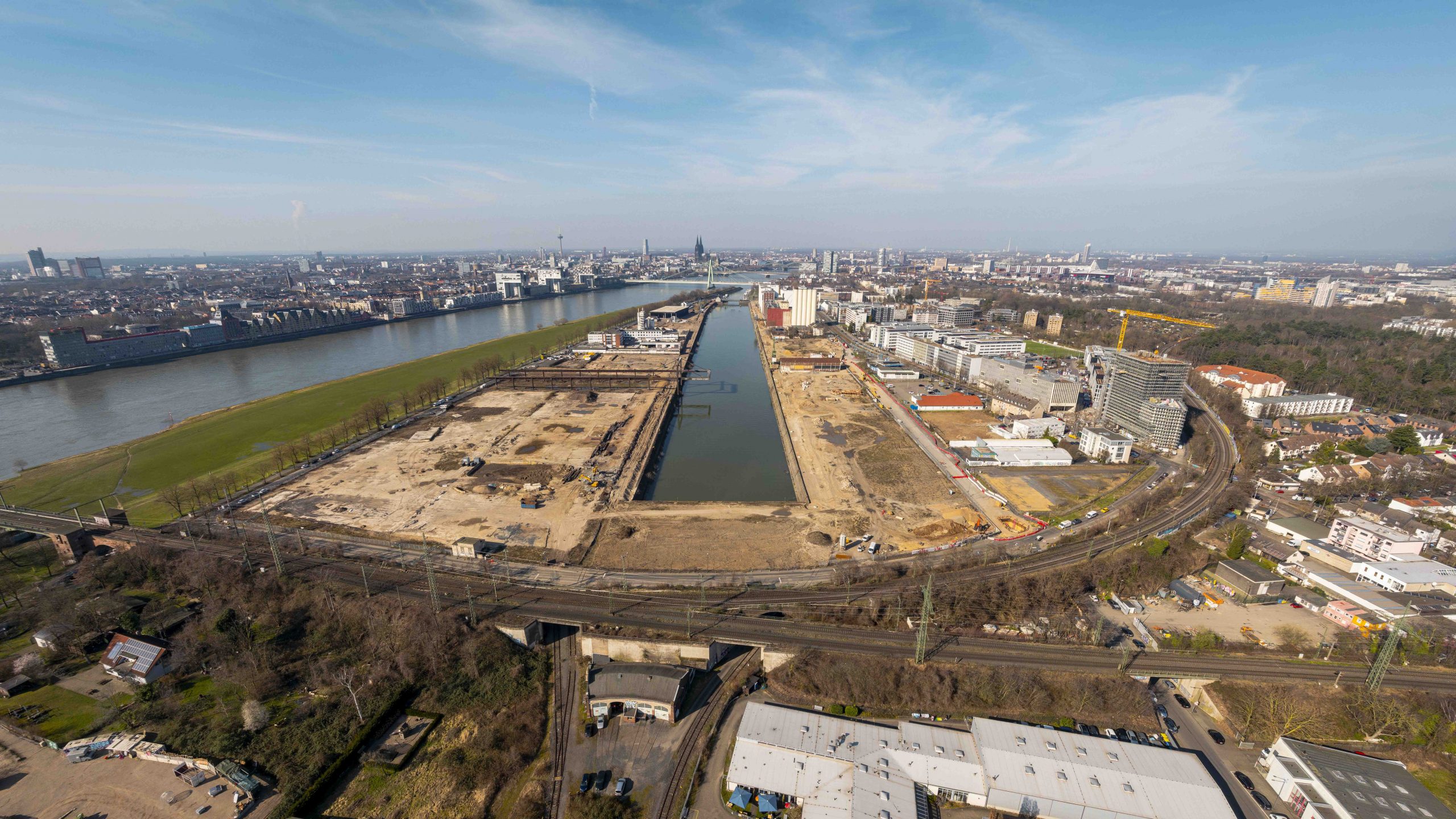 Luftaufnahme des gesamten Deutzer Hafens in Köln: Der Hafenkanal verläuft mittig durch das Areal, links liegt der Rhein mit Grünstreifen und gegenüberliegender Stadtsilhouette. Auf beiden Seiten des Hafenbeckens sind große Baustellenflächen, Bestandsgebäude und erste Neubauten zu sehen. Im Vordergrund verlaufen Bahntrassen und Straßen, im Hintergrund die Kölner Innenstadt mit Dom.