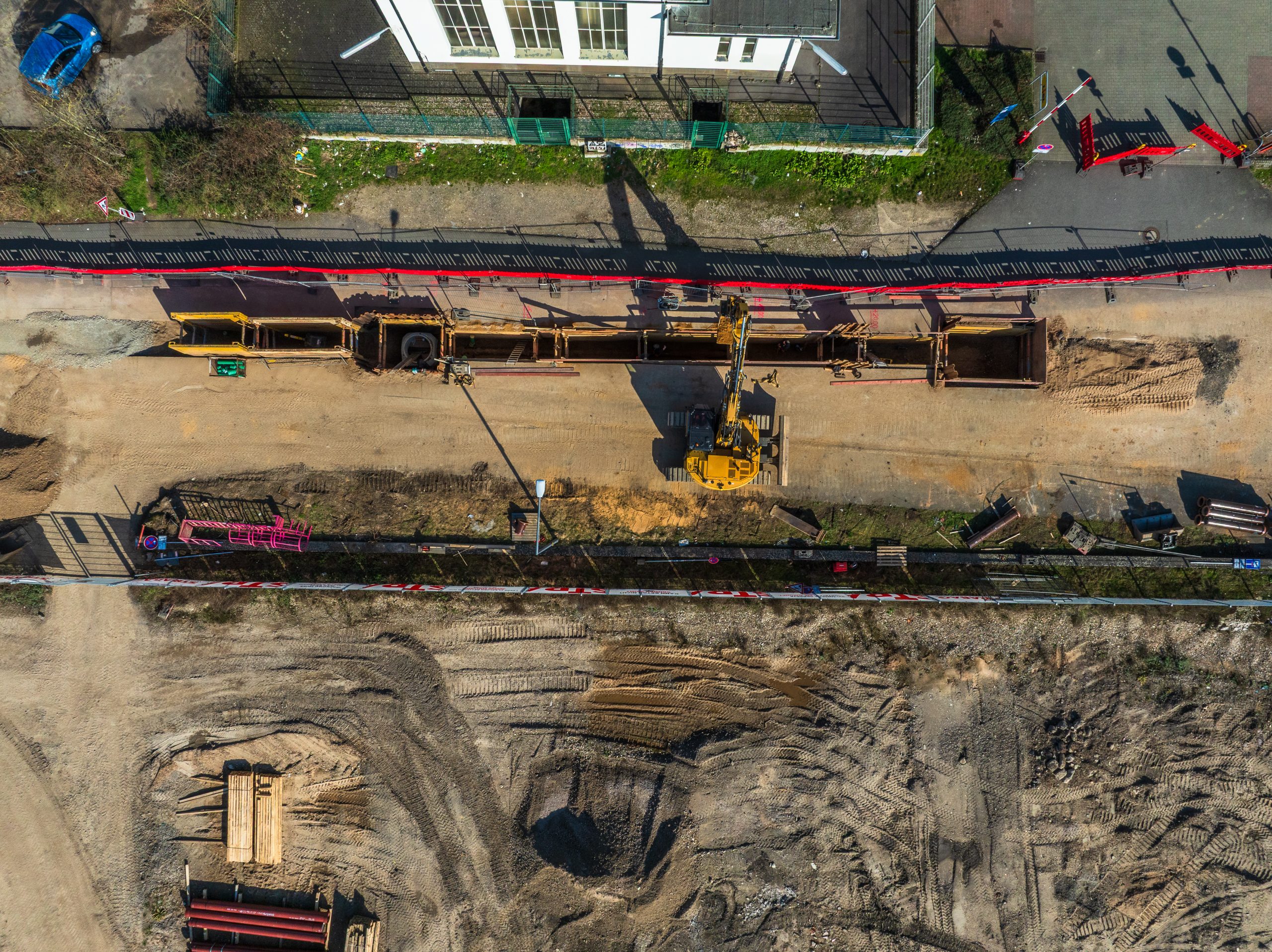 Draufsicht auf Kanalbauarbeiten im Deutzer Hafen auf der Ostseite: Ein Bagger arbeitet entlang eines geöffneten Leitungsgrabens mit verbauter Baugrube. Umgeben von Baustellenflächen, Absperrungen und gelagerten Rohren werden Versorgungsleitungen im Boden verlegt.