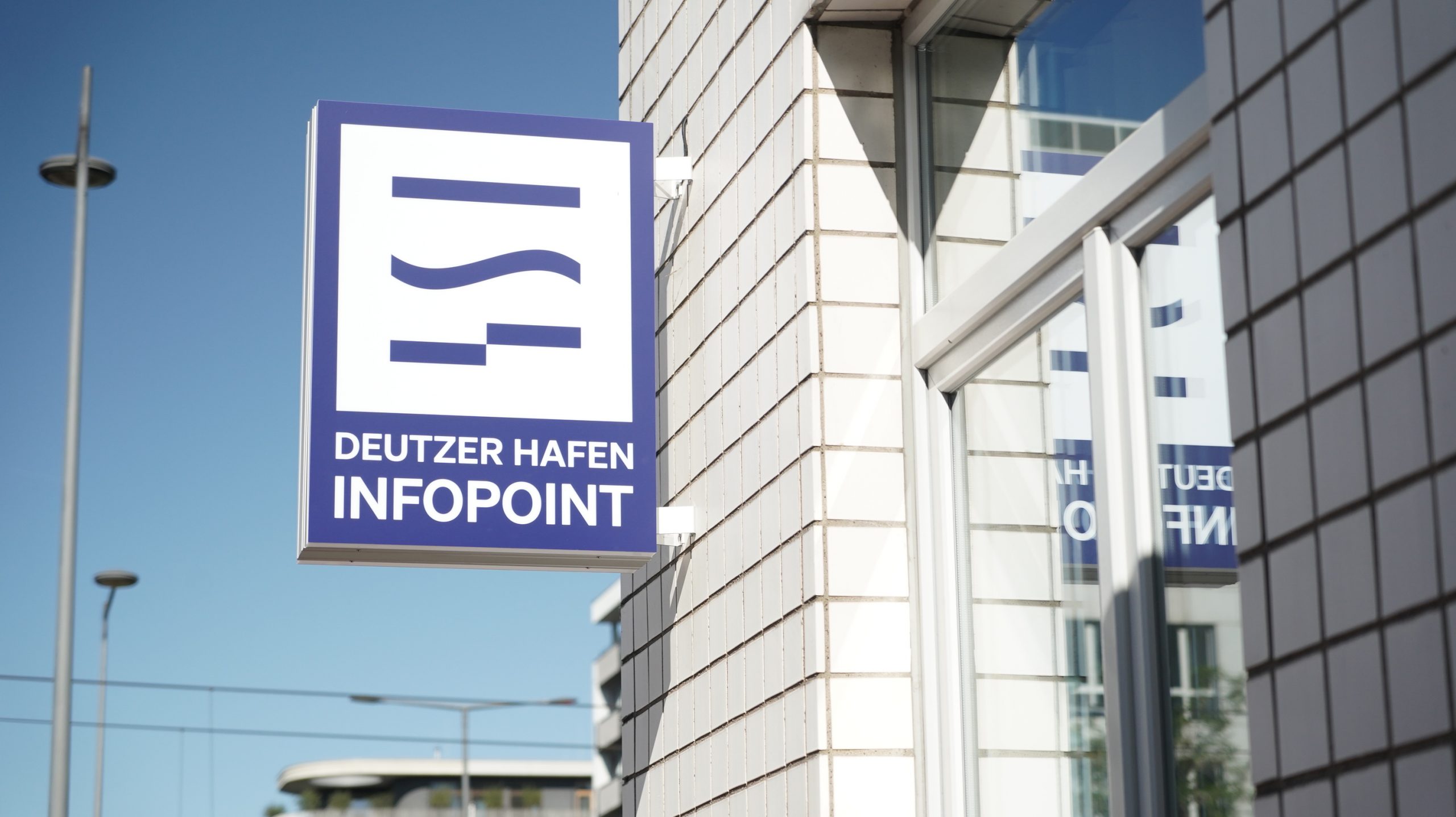 Außenschild des Infopoints Deutzer Hafen Köln. Ein blau-weißes quadratisches Schild mit dem Logo und Schriftzug „Deutzer Hafen Infopoint" ist an einer modernen Glasfassade montiert. Der Infopoint ist die zentrale Anlaufstelle für Informationen rund um das Stadtentwicklungsprojekt Deutzer Hafen Köln.