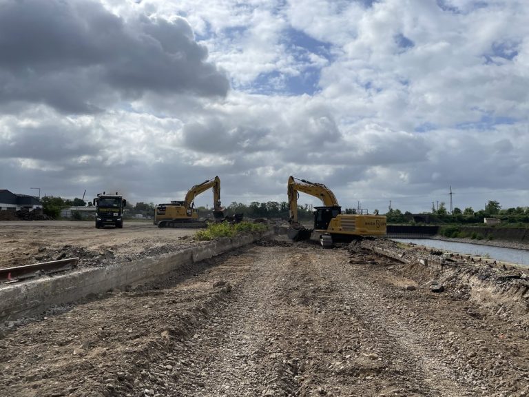 Bauarbeiten auf dem Gelände des Deutzer Hafens in Köln. Zwei gelbe Kettenbagger arbeiten nebeneinander beim Abbruch alter Bahngleisanlagen und Betonfundamente. Ein Lkw steht links bereit für den Abtransport des Abbruchmaterials. Im Hintergrund rechts ist das Hafenbecken erkennbar. Schotter, Betonbruchstücke und freiliegende Gleisreste bedecken den Untergrund im Vordergrund. Bewölkter Sommerhimmel über der weitläufigen Baustelle.
