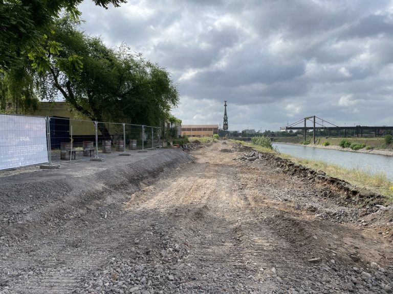 Bodennahe Aufnahme des geräumten Uferbereichs am Hafenbecken des Deutzer Hafens in Köln. Im Vordergrund verläuft ein aufgeschütteter Erdweg entlang der Kaimauer. Links steht ein Bauzaun mit Projektinformationstafel sowie ein großer Laubbaum. Im Hintergrund ist der historische grüne Portalkran am Hafenbecken erkennbar, dahinter die rostbraune Stahlkonstruktion der alten Verladebrücke. Rechts liegt das Hafenbecken. Bewölkter Sommerhimmel über der Baustelle.