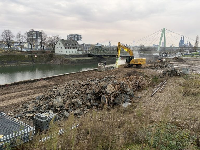 ALT-Text (Kaimauer-Rückbau am Hafenbecken) Abbrucharbeiten an der Kaimauer des Deutzer Hafens in Köln. Ein gelber Kettenbagger bricht die Betonkaimauer am Hafenbecken ab, Staub steigt beim Abbruch auf. Im Vordergrund liegt ein großer Haufen Betonbruchstücke mit freiliegenden Bewehrungsstählen sowie abgelagerte Stahlrohre. Im Hintergrund sind die historische grüne Drehbrücke am Hafeneingang, ein weißes Wohnhaus sowie die Severinsbrücke und der Kölner Dom erkennbar. Links liegt das Hafenbecken, dahinter die Poller Wiese mit kahlen Winterbäumen.