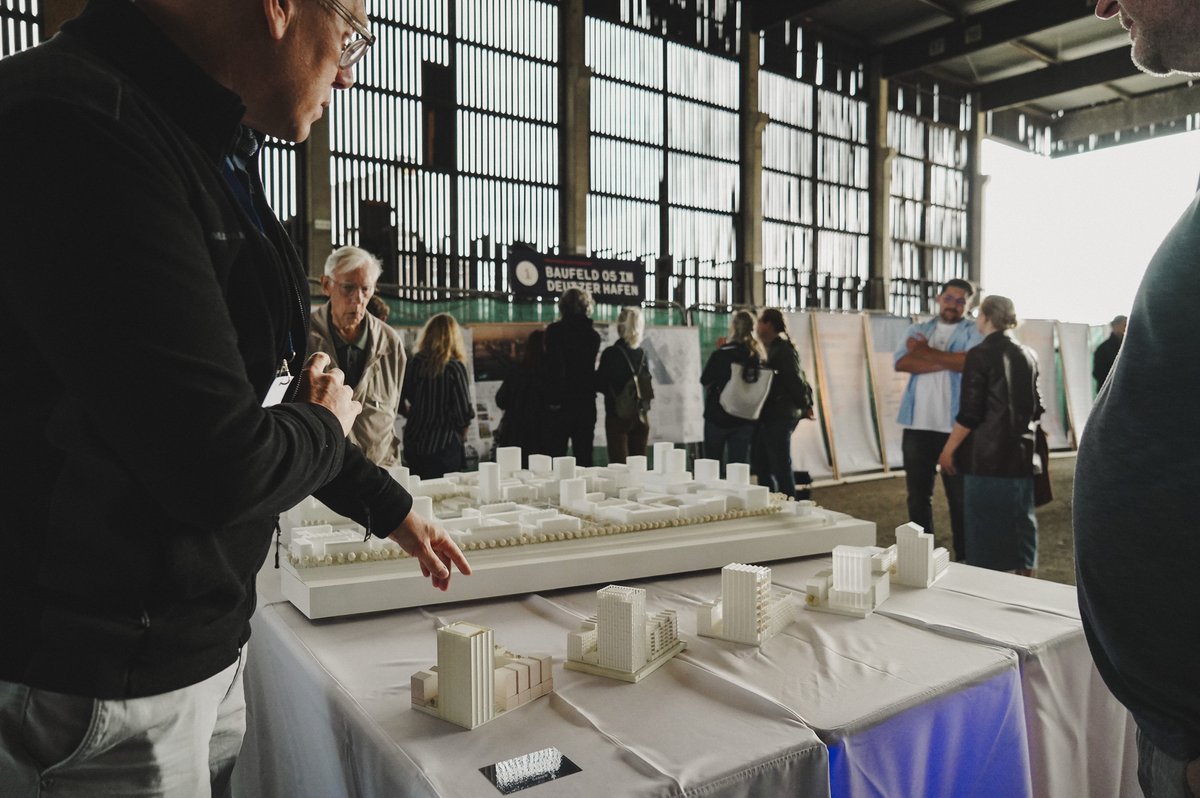 Menschen betrachten Architekturmodelle auf Tischen in einer großen Industriehalle. Mehrere Besucher stehen um die ausgestellten Stadt- und Gebäudeentwürfe, während im Hintergrund weitere Personen miteinander ins Gespräch vertieft sind.