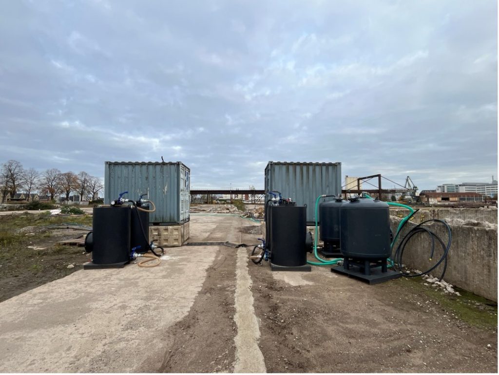 ALT-Text (Air-Sparging-Anlage / Deutzer Hafen) Bodensanierungsanlage auf dem Baugelände des Deutzer Hafens in Köln. Zwei Baucontainer mit je mehreren schwarzen Drucktanks und grünen Schlauchleitungen stehen auf einer Betonplatte — Bestandteile einer Air-Sparging-Anlage zur Grundwassersanierung. Im Hintergrund sind die geräumte Brachfläche, die rostbraune Stahlkonstruktion der alten Verladebrücke sowie der grüne Portalkran am Hafenbecken erkennbar. Bewölkter Winterhimmel über dem Gelände.