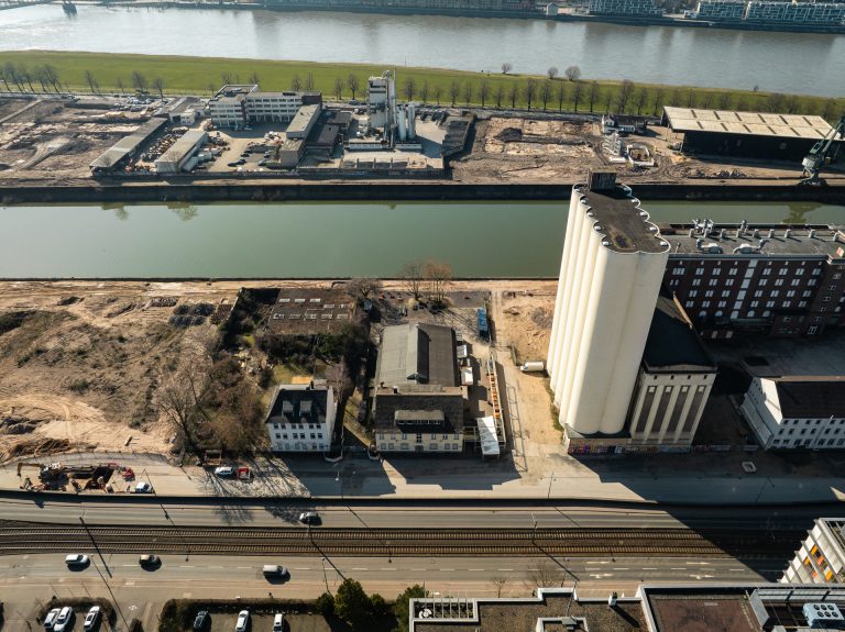 Drohnenaufnahme der Zwischenzone zwischen Ellmühle und Auermühle am Deutzer Hafen. Im Vordergrund ältere Bestandsgebäude, die noch auf dem Baufeld stehen. Rechts dominieren die weißen zylindrischen Silos. Über das Hafenbecken ist die Hafenzunge mit Gewerbebebauung, der Alfred-Schütte-Allee und der Poller Wiese zu sehen. Im Hintergrund der Rhein.