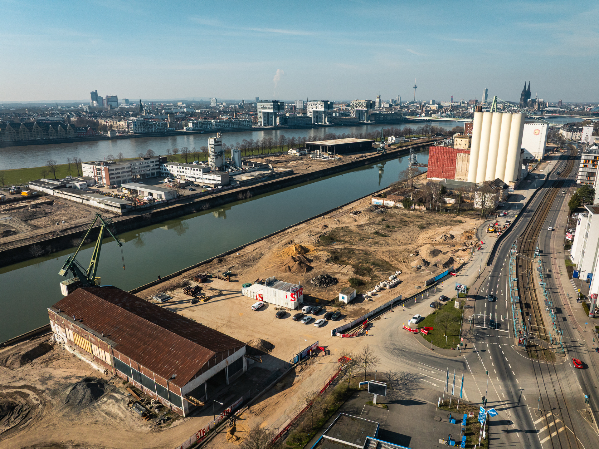 Luftaufnahme des südöstlichen Baufelds am Deutzer Hafen. Im Vordergrund ein rot-brauner Backsteinlagergebäude sowie Baucontainer der Firma Strabag. Rechts die weißen Silos der Ellmühle. Das Hafenbecken spiegelt die Baukräne. Im Hintergrund über den Rhein die Kölner Innenstadt mit Dom, Colonius und Severinsbrücke.