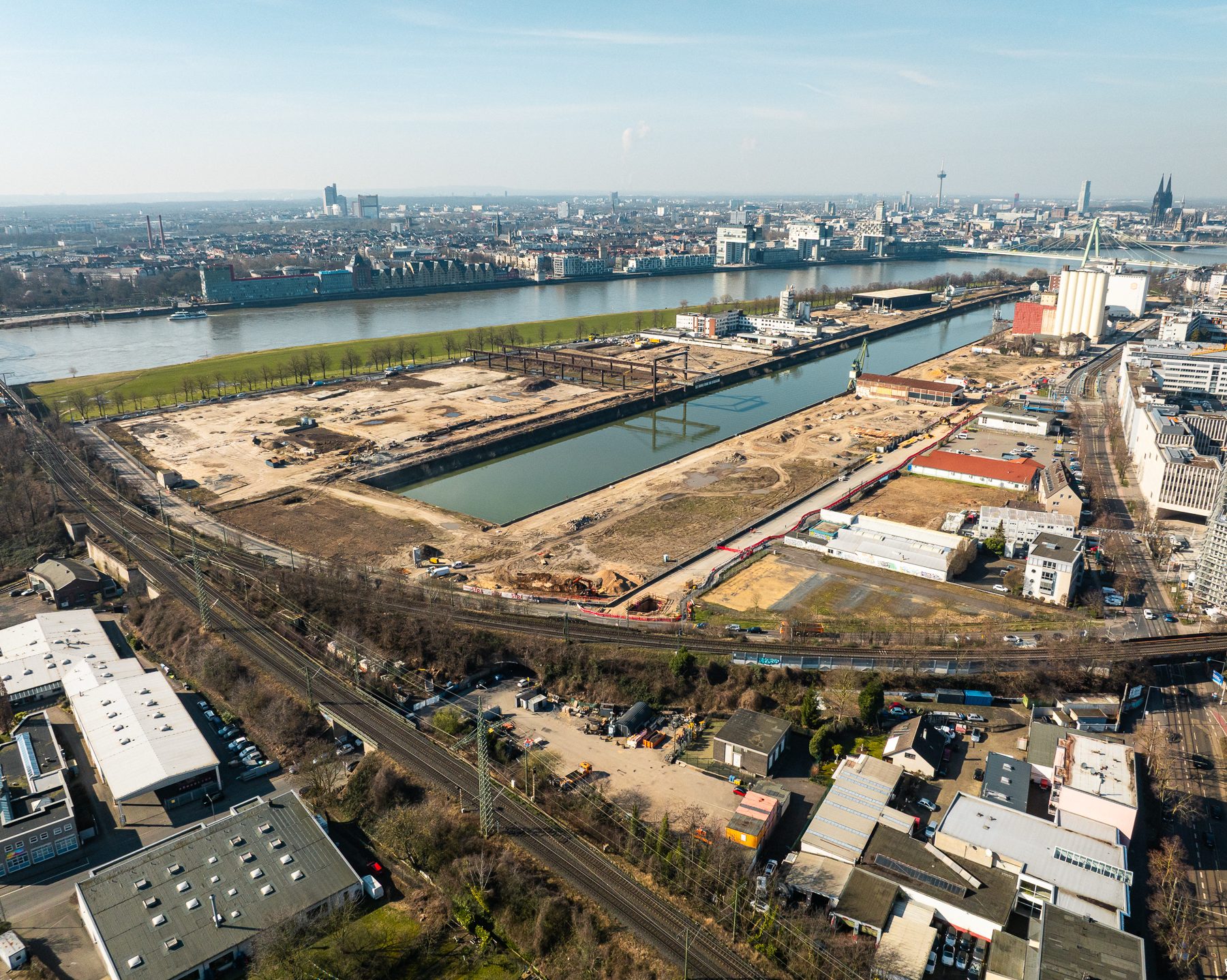 Weiträumige Luftaufnahme des gesamten Deutzer Hafens aus südlicher Perspektive. Das Hafenbecken teilt das Bild. Links das ausgedehnte Baufeld mit dem Portalkran-Kunstwerk. Rechts weitere Bauflächen mit Baucontainern und Kränen. Im Vordergrund Gewerbebebauung, Gleisanlagen und Bahntrassen. Im Hintergrund der Rhein mit Severinsbrücke und der Kölner Skyline mit Dom.