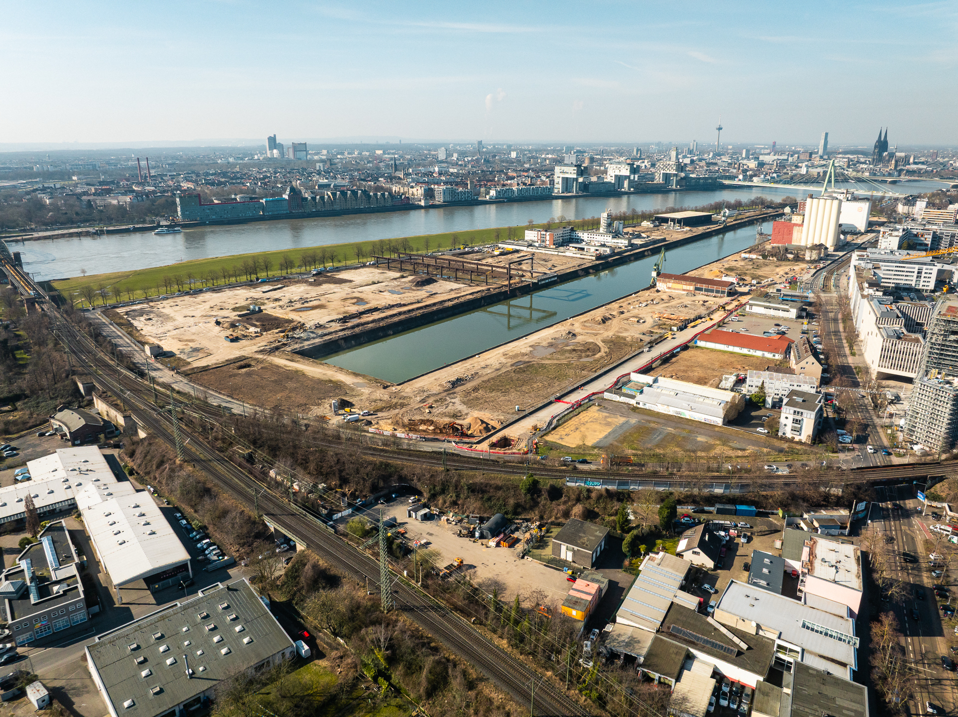 Weiträumige Luftaufnahme des gesamten Deutzer Hafens aus südlicher Perspektive. Das Hafenbecken teilt das Bild. Links das ausgedehnte Baufeld mit dem Portalkran-Kunstwerk. Rechts weitere Bauflächen mit Baucontainern und Kränen. Im Vordergrund Gewerbebebauung, Gleisanlagen und Bahntrassen. Im Hintergrund der Rhein mit Severinsbrücke und der Kölner Skyline mit Dom.