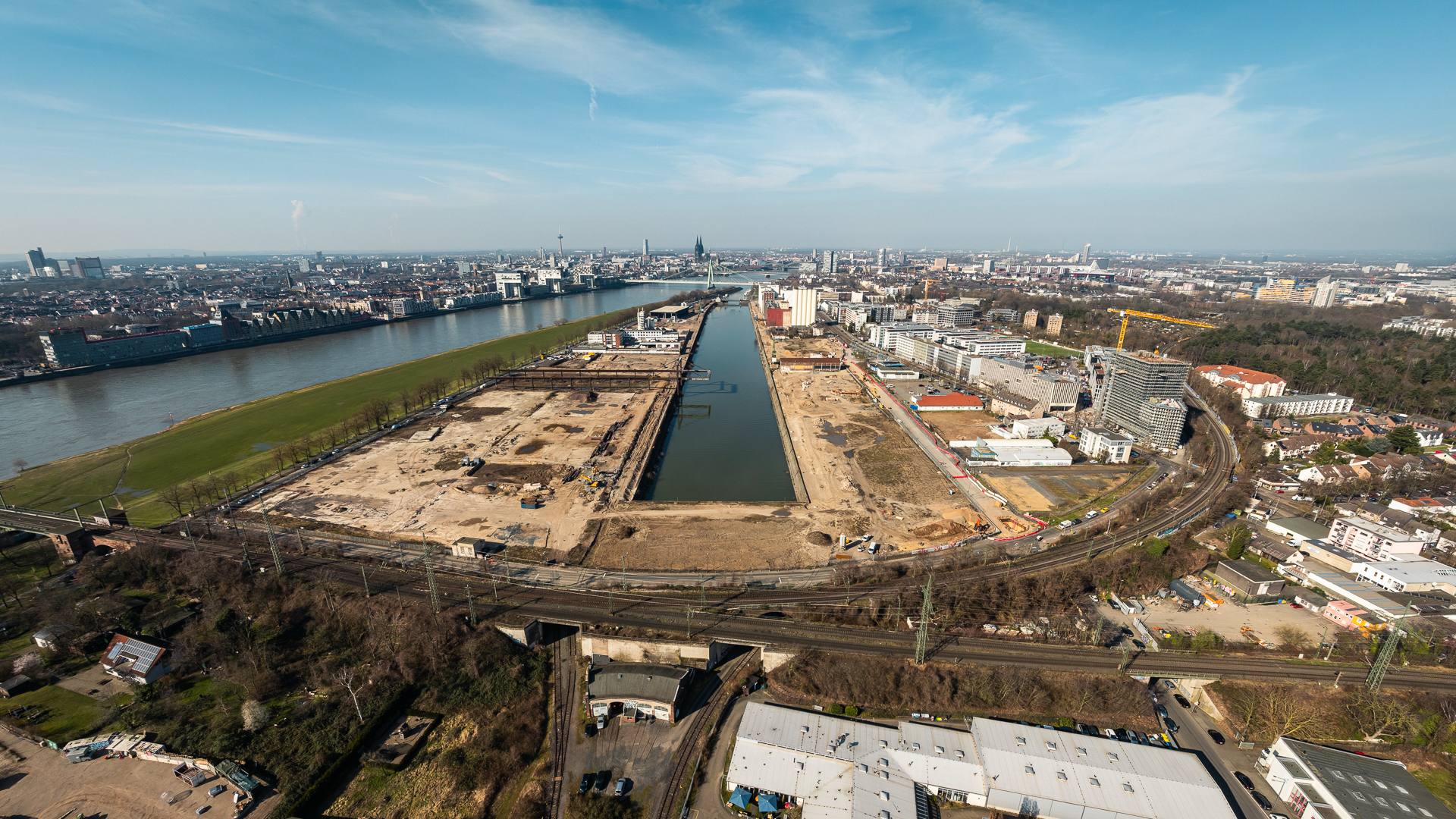 Großformatiges Drohnenpanorama des Deutzer Hafens aus großer Höhe. Das gesamte Hafenareal mit dem langgestreckten Hafenbecken ist in der Bildmitte erkennbar. Links die Poller Wiese am Rhein, rechts die Stadtlandschaft von Köln-Deutz mit Neubebauung und LVR-Turm. Im Vordergrund Gewerbeflächen und Gleisanlagen. Im Hintergrund der Rhein und die Kölner Innenstadt mit Dom und Colonius.
