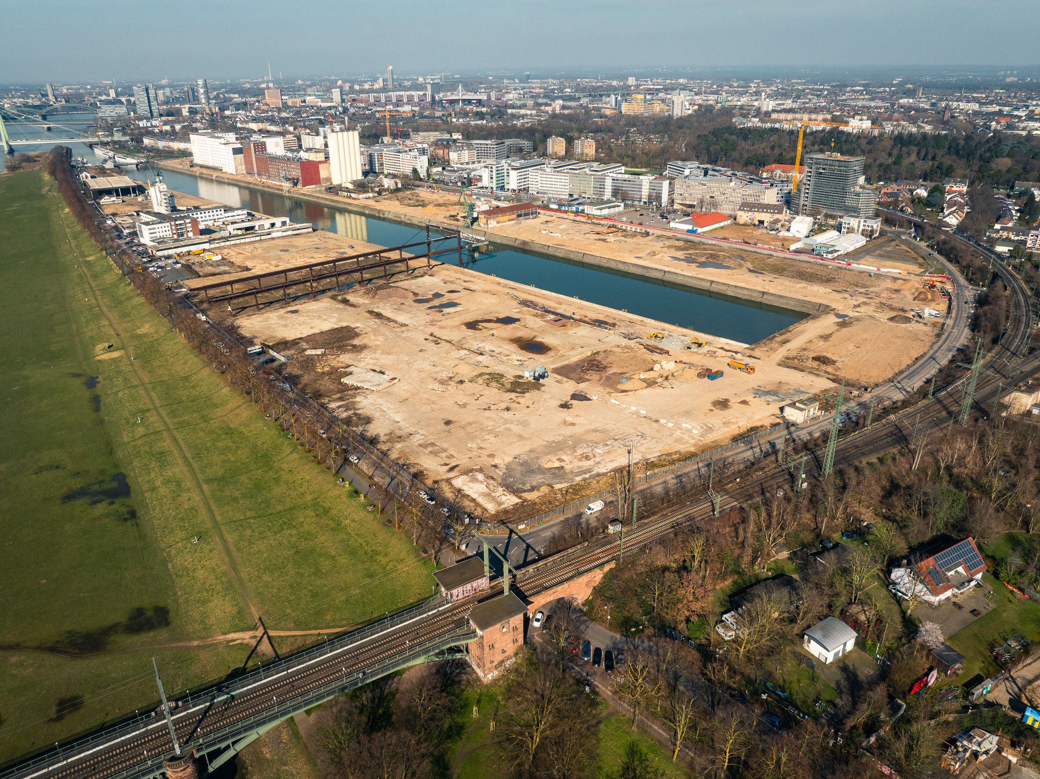 Luftaufnahme der Baustelle am Deutzer Hafen in Köln. Im Vordergrund verläuft eine Eisenbahnbrücke über die Poller Wiese. Dahinter liegt das weitgehend geräumte, sandige Baufeld, das vom Hafenbecken begrenzt wird. Am linken Rand sind Reste einer alten Stahlkonstruktion zu sehen. Mehrere Baufahrzeuge sind auf dem Gelände im Einsatz. Im Hintergrund erstreckt sich die Kölner Stadtsilhouette mit Bürogebäuden und Wohnhäusern. Rechts verläuft die Alfred-Schütte-Allee mit einer geschwungenen Straßenführung entlang des Hafenbeckens.