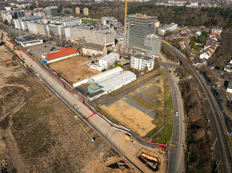 Luftaufnahme der Baustelle am Deutzer Hafen. Im Zentrum des Bildes sind noch bestehende Gewerbegebäude sowie eine Halle mit Graffiti zu sehen, die von der Baustelle umgeben sind. Entlang der Alfred-Schütte-Allee (rechts) markiert eine rote Absperrung die Baustellengrenze. Im Hintergrund ist ein im Rohbau befindliches Hochhaus mit Baukran erkennbar. Im Vordergrund befindet sich eine Baugrube mit Holzverschalung.