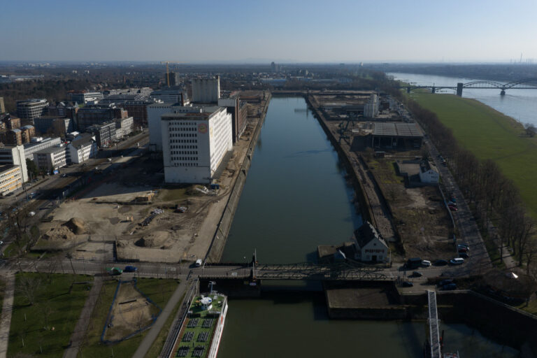 Drohnenaufnahme entlang des Hafenbeckens am Deutzer Hafen, Blickrichtung Rhein. Links am Ufer steht die weiße Ellmühle mit Baugerüsten, rechts die Hafenzunge mit einem Verwaltungsgebäude und einer Lagerhalle. Im Hintergrund mündet das Hafenbecken in den Rhein, über den die Rodenkirchener Brücke führt. Rechts oben erstreckt sich die grüne Poller Wiese.