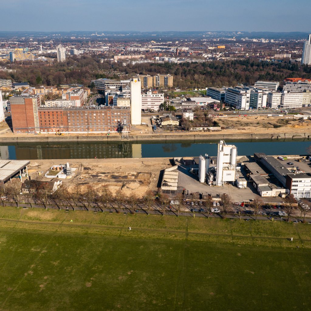 Luftaufnahme des Deutzer Hafens vom Rheinufer aus. Im Vordergrund die grüne Poller Wiese, dahinter die Alfred-Schütte-Allee mit parkenden Fahrzeugen. Auf dem Areal der STRABAG sind ein weißer Industriesilo sowie Lagerhallen zu erkennen. Gegenüber auf der anderen Seite des Hafenbeckens spiegeln sich Ellmühle und Auermühle im Wasser. Im Hintergrund die Büro- und Gewerbebebauung von Köln-Deutz.