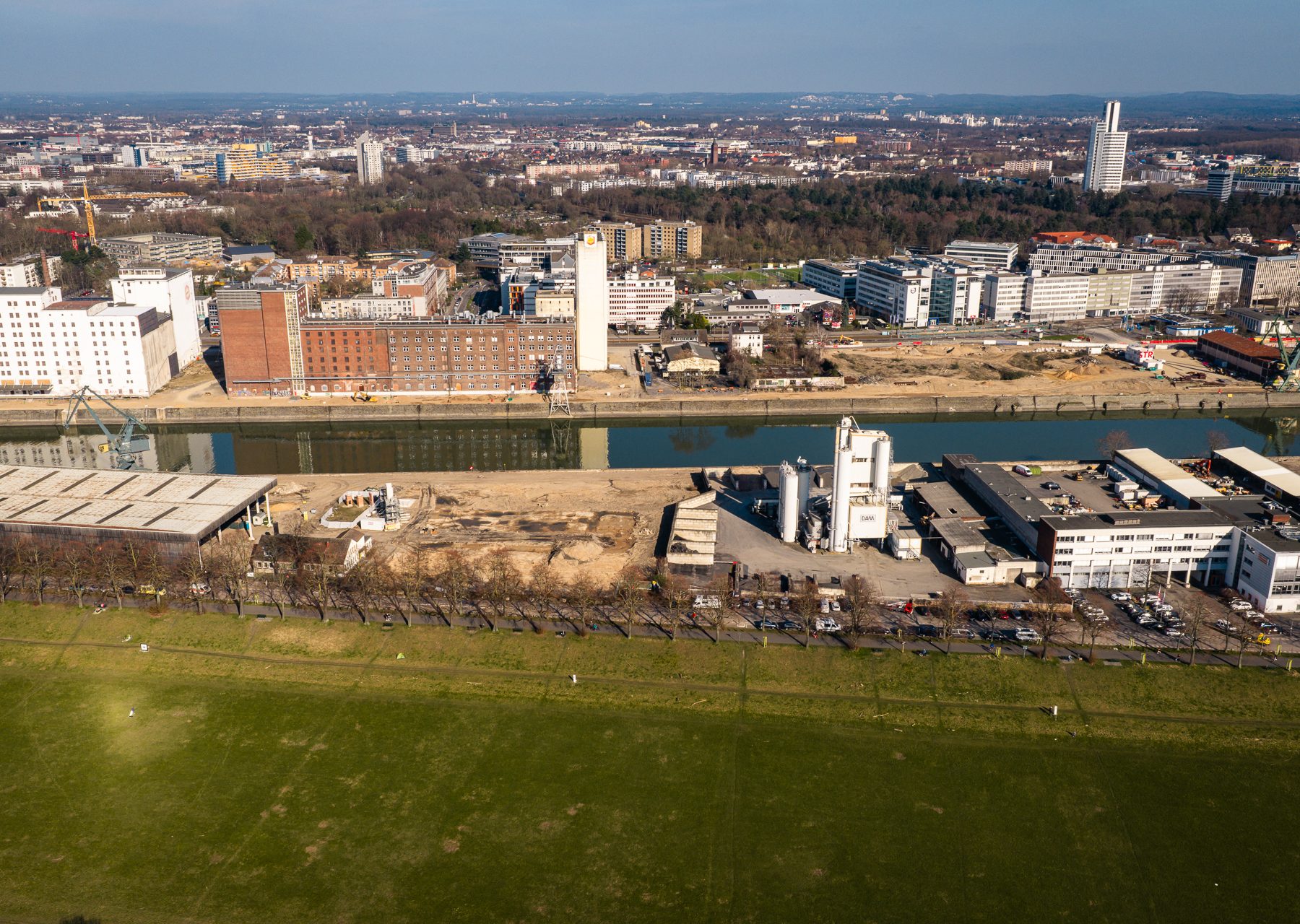 Luftaufnahme des Deutzer Hafens vom Rheinufer aus. Im Vordergrund die grüne Poller Wiese, dahinter die Alfred-Schütte-Allee mit parkenden Fahrzeugen. Auf dem Areal der STRABAG sind ein weißer Industriesilo sowie Lagerhallen zu erkennen. Gegenüber auf der anderen Seite des Hafenbeckens spiegeln sich Ellmühle und Auermühle im Wasser. Im Hintergrund die Büro- und Gewerbebebauung von Köln-Deutz.