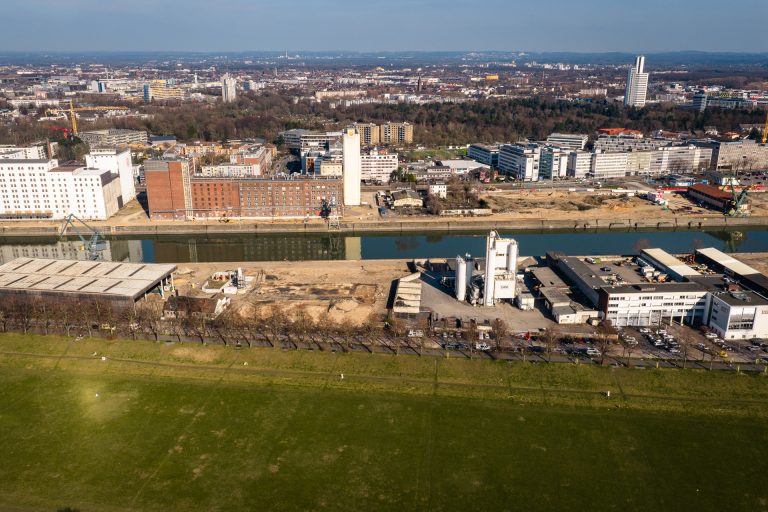 Luftaufnahme des Deutzer Hafens vom Rheinufer aus. Im Vordergrund die grüne Poller Wiese, dahinter die Alfred-Schütte-Allee mit parkenden Fahrzeugen. Auf dem Areal der STRABAG sind ein weißer Industriesilo sowie Lagerhallen zu erkennen. Gegenüber auf der anderen Seite des Hafenbeckens spiegeln sich Ellmühle und Auermühle im Wasser. Im Hintergrund die Büro- und Gewerbebebauung von Köln-Deutz.