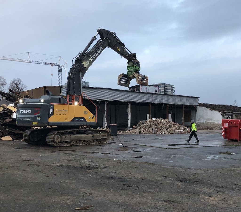 Bagger mit Greifarm beim Abriss einer Industriehalle im Deutzer Hafen Köln, daneben Schuttberge und eine Person in Warnkleidung.