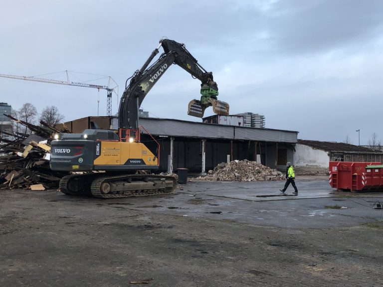 Bagger mit Greifarm beim Abriss einer Industriehalle im Deutzer Hafen Köln, daneben Schuttberge und eine Person in Warnkleidung.