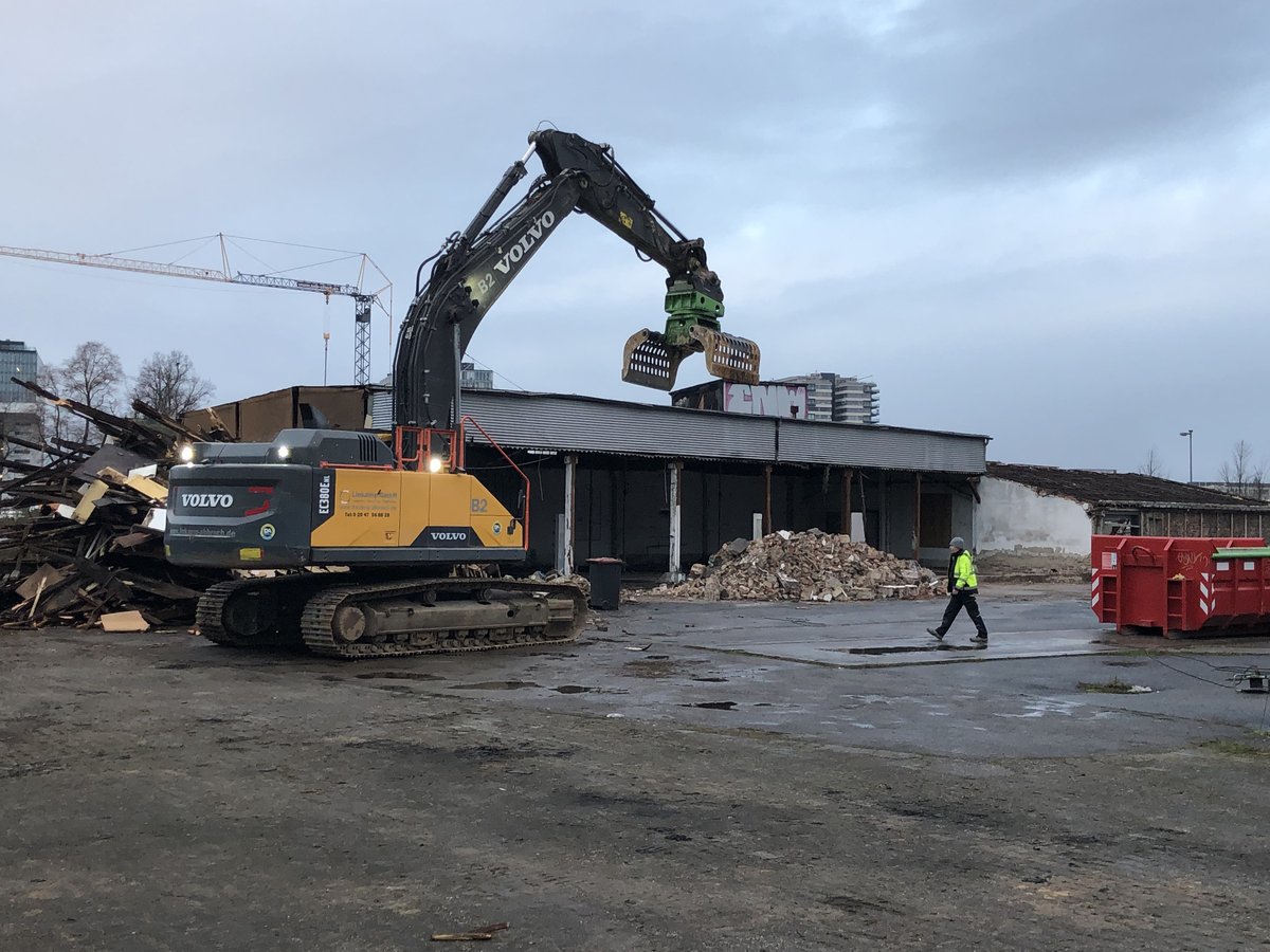 Bagger mit Greifarm beim Abriss einer Industriehalle im Deutzer Hafen Köln, daneben Schuttberge und eine Person in Warnkleidung.