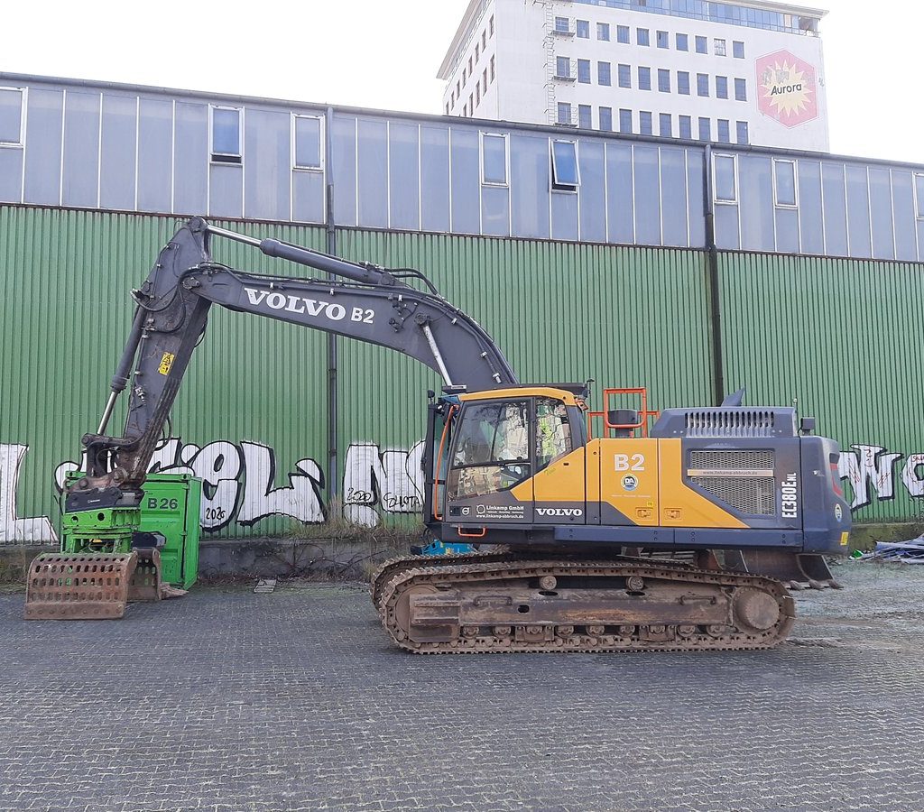 Ein großer Kettenbagger steht auf einem gepflasterten Industriegelände im Deutzer Hafen Köln vor einer langen grünen Lagerhalle mit Graffiti. Der Bagger hat einen Greifaufsatz und ist seitlich ausgerichtet. Im Hintergrund ragt ein höheres Bürogebäude mit „Aurora“-Logo über die Halle hinaus.