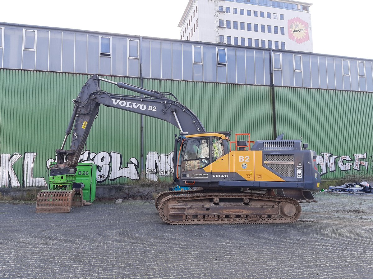 Ein großer Kettenbagger steht auf einem gepflasterten Industriegelände im Deutzer Hafen Köln vor einer langen grünen Lagerhalle mit Graffiti. Der Bagger hat einen Greifaufsatz und ist seitlich ausgerichtet. Im Hintergrund ragt ein höheres Bürogebäude mit „Aurora“-Logo über die Halle hinaus.