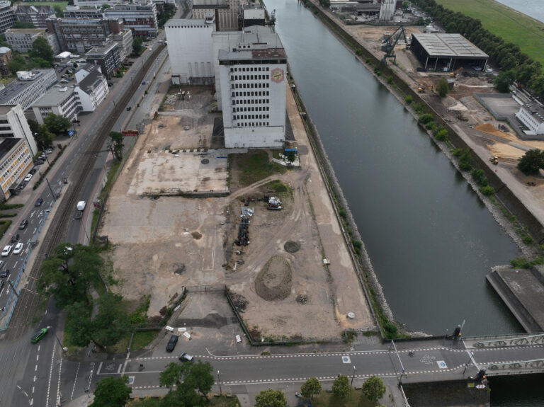 Luftaufnahme des Deutzer Hafens Köln mit Blick entlang des Hafenbeckens Richtung Norden. Links das Aurora-Hochhaus der Ellmühle und die Alfred-Schütte-Allee. Rechts ist auf der gegenüberliegenden Uferseite reger Baubetrieb mit Baggern und Materiallagern erkennbar, dahinter die Poller Wiese mit Baumreihe entlang des Rheins.