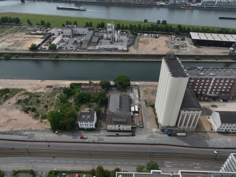 Luftaufnahme der Auermühle und der Siloanlage am Deutzer Hafen Köln. Die hohen weißen Betonsilos ragen über das Hafenbecken, daneben das rote Backsteingebäude. Im Vordergrund liegt brachliegendes, teils begrüntes Gelände, im Hintergrund der Rhein mit Frachtschiffen und die Kölner Innenstadt.