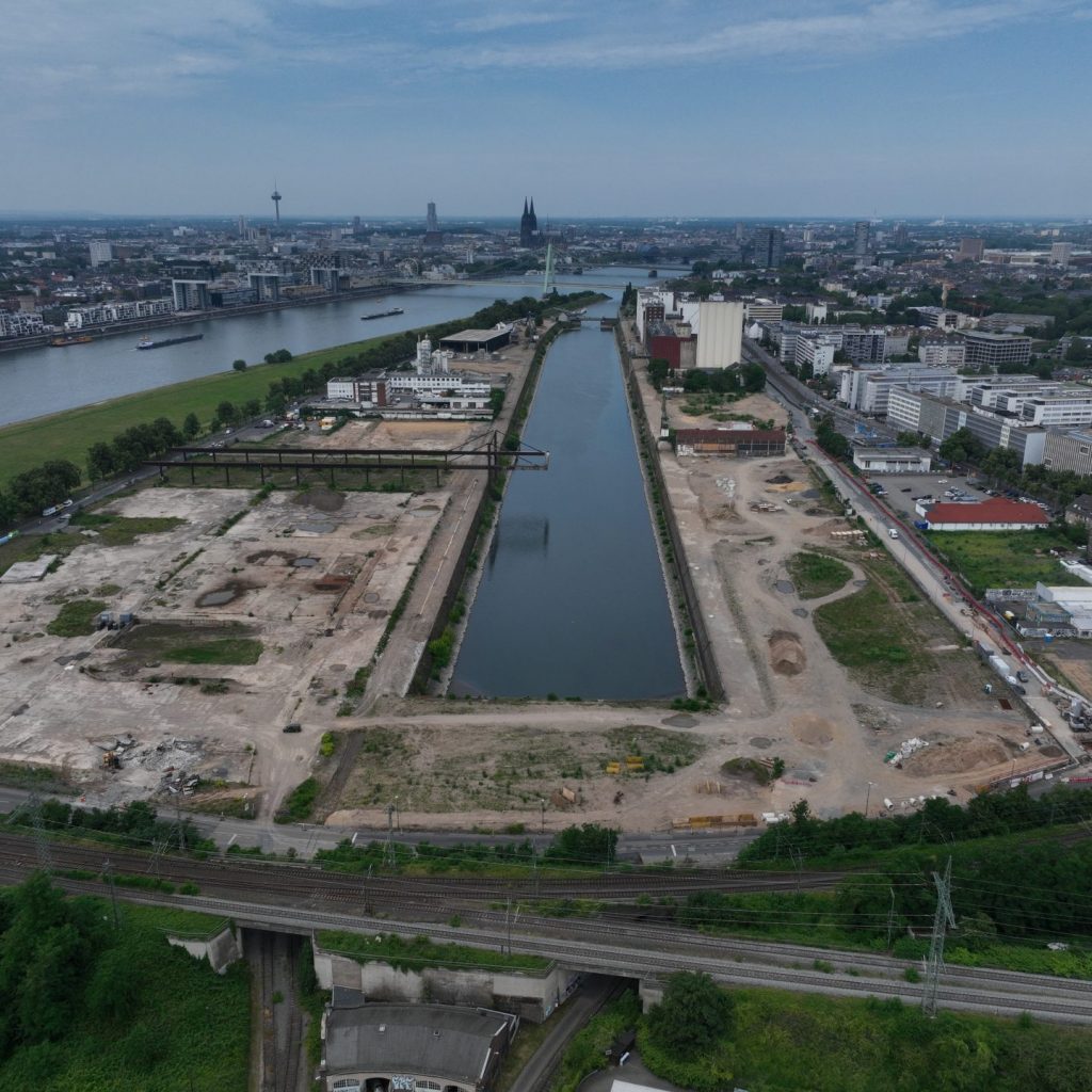 Drohnenaufnahme der Gesamtfläche am Deutzer Hafen mit langem Hafenbecken in der Bildmitte, umgeben von freigeräumten Brachflächen. Links die Poller Wiese am Rhein, im Hintergrund die Severinsbrücke und die Kölner Altstadt mit Dom.