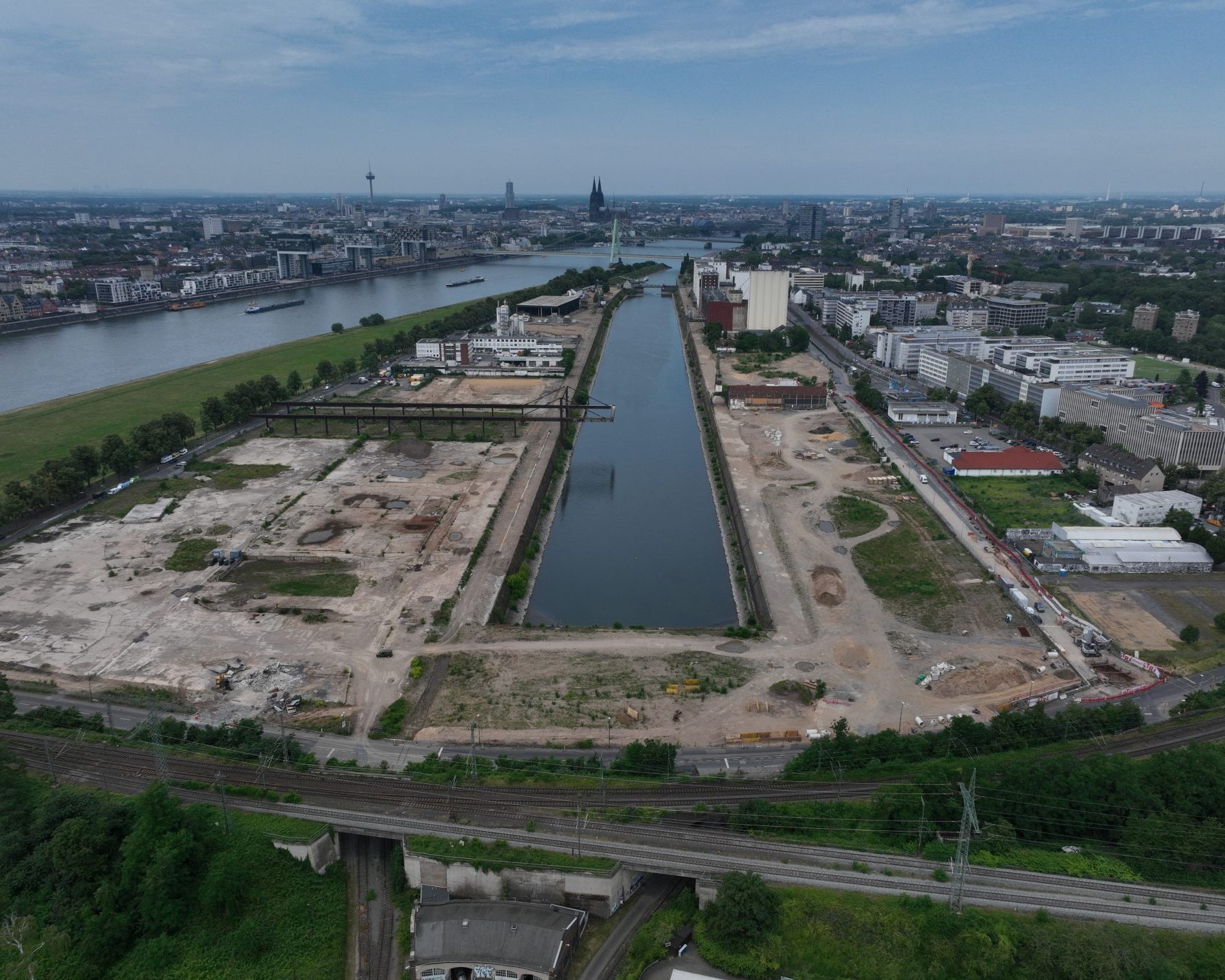 Drohnenaufnahme der Gesamtfläche am Deutzer Hafen mit langem Hafenbecken in der Bildmitte, umgeben von freigeräumten Brachflächen. Links die Poller Wiese am Rhein, im Hintergrund die Severinsbrücke und die Kölner Altstadt mit Dom.