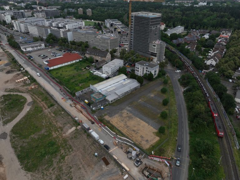 Luftaufnahme eines eingerüsteten Hochhauses im Bau an der Siegburger Straße, umgeben von Bürogebäuden, einer Lagerhalle mit Graffiti sowie Bahngleisen.