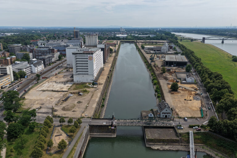 Drohnenaufnahme entlang des Hafenbeckens am Deutzer Hafen, Blickrichtung Rhein. Links am Ufer steht die weiße Ellmühle mit Baugerüsten, rechts die Hafenzunge mit einem Verwaltungsgebäude und einer Lagerhalle. Im Hintergrund mündet das Hafenbecken in den Rhein, über den die Rodenkirchener Brücke führt. Rechts oben erstreckt sich die grüne Poller Wiese.