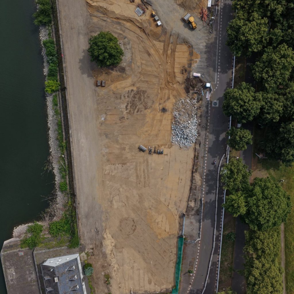 Senkrechte Luftaufnahme der Baustelle Deutzer Hafen Köln entlang des Hafenbeckens. Zu sehen sind Erdarbeiten mit Baufahrzeugen, gelagertem Baumaterial und Bauschutt sowie ein Bestandsgebäude mit hellblauer Fassade. Rechts verläuft ein Rad- und Fußweg mit Baumreihen entlang der Poller Wiese.