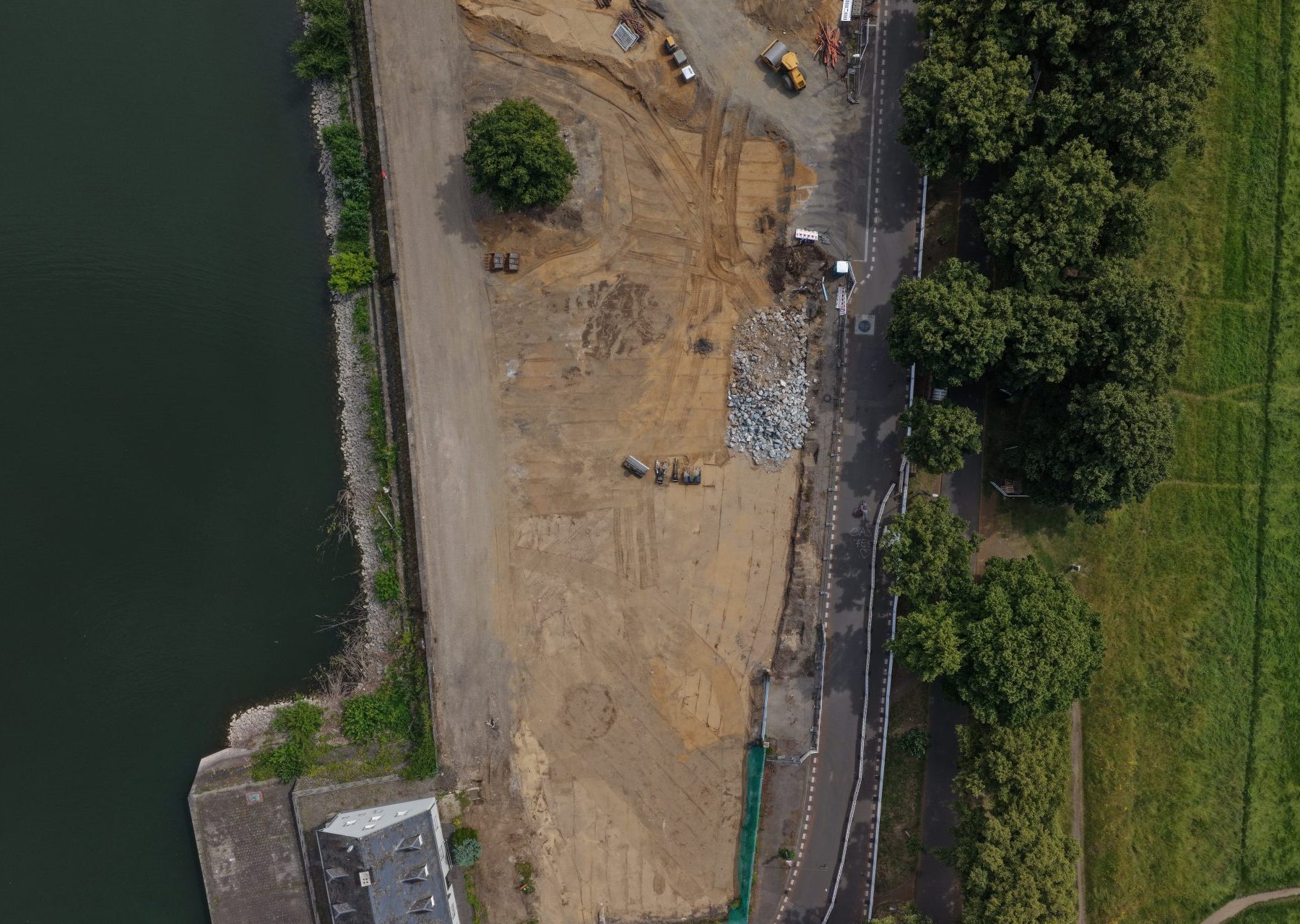 Senkrechte Luftaufnahme der Baustelle Deutzer Hafen Köln entlang des Hafenbeckens. Zu sehen sind Erdarbeiten mit Baufahrzeugen, gelagertem Baumaterial und Bauschutt sowie ein Bestandsgebäude mit hellblauer Fassade. Rechts verläuft ein Rad- und Fußweg mit Baumreihen entlang der Poller Wiese.