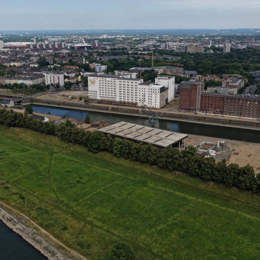 Luftaufnahme der Baustelle Deutzer Hafen Köln mit Blick auf das Hafenbecken. Links die weiße Ellmühle und die backsteinrote Auermühle, davor Kräne und Erschließungsflächen. Rechts ein Betriebsgelände mit weißen Silotürmen, im Hintergrund die Poller Wiese sowie die Deutzer Stadtsilhouette.
