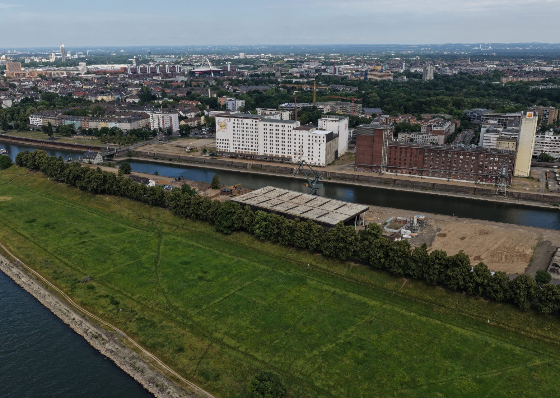 Luftaufnahme der Baustelle Deutzer Hafen Köln mit Blick auf das Hafenbecken. Links die weiße Ellmühle und die backsteinrote Auermühle, davor Kräne und Erschließungsflächen. Rechts ein Betriebsgelände mit weißen Silotürmen, im Hintergrund die Poller Wiese sowie die Deutzer Stadtsilhouette.