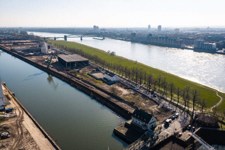 Luftaufnahme der Baustelle Deutzer Hafen Köln mit Blick über das Hafenbecken und den Rhein auf die Kölner Rheinauhafen-Silhouette mit den Gewölben der Hafenspeicher, aufgenommen im März. Im Vordergrund die Holzhalle sowie das Gebäude der Wasserschutzpolizei, dazwischen die Poller Wiese mit kahlen Baumreihen. Im Hintergrund eine Bogenbrücke über den Rhein.