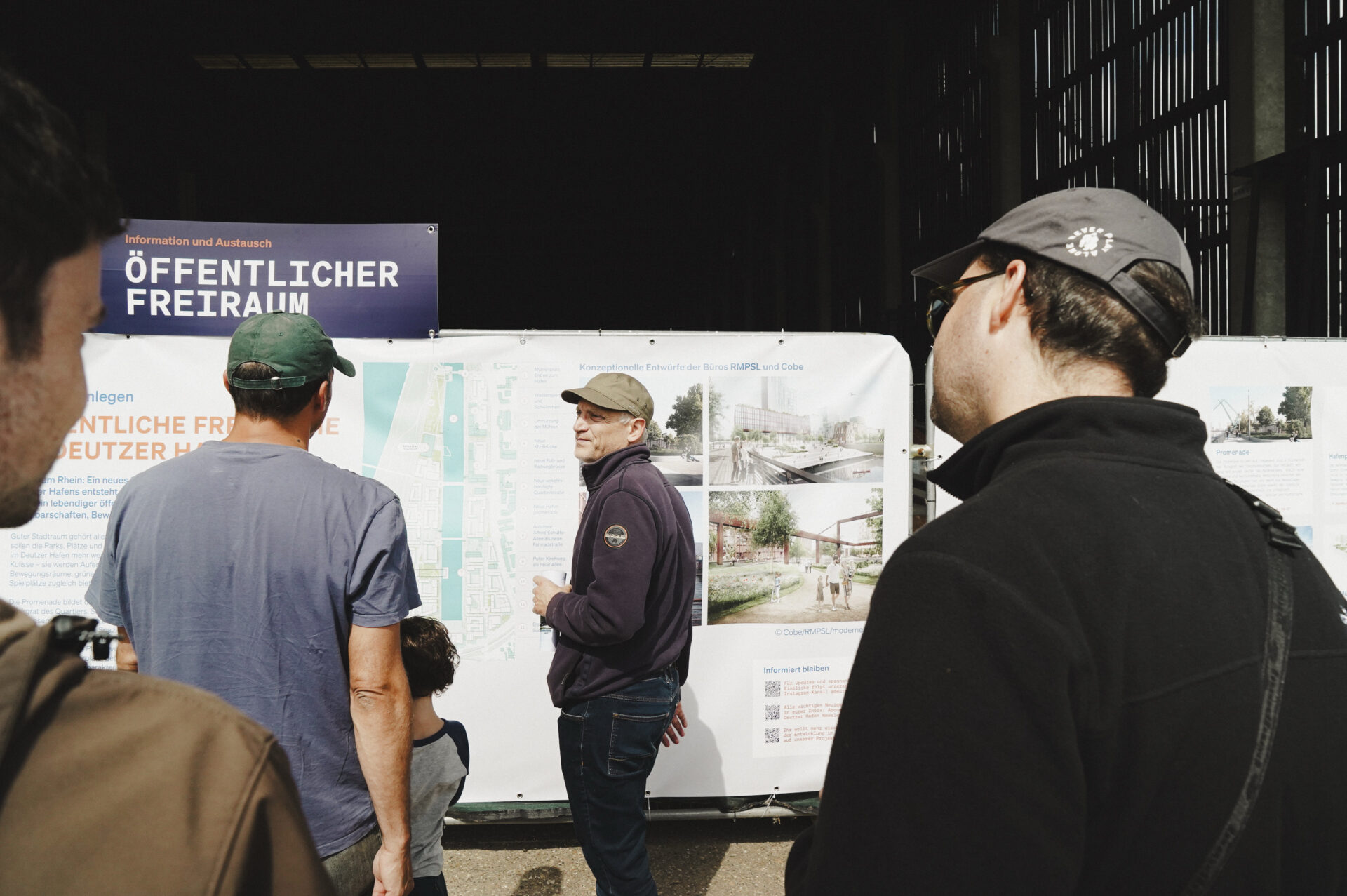 Gruppe von Personen steht vor einem Informationsstand mit Plakaten über den Hafentag 2025. Sichtbare Texte beinhalten 'ÖFFENTLICHER FREIRAUM' und Pläne für zukünftige Entwicklungen.