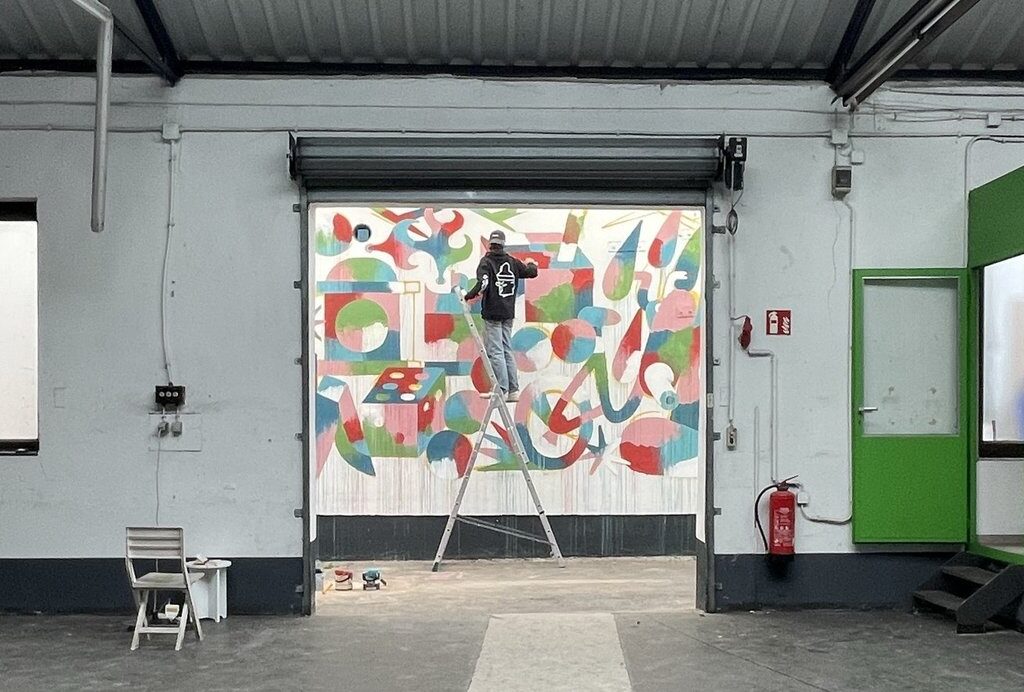 Ein Mann steht in einer Industriehalle auf einer Leiter. Er malt ein Graffitti an eine Wand.